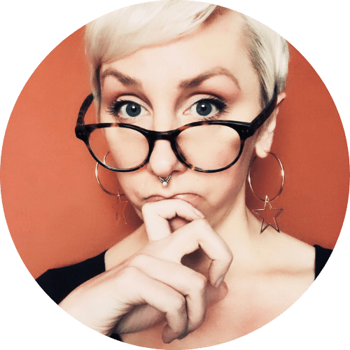 Photo of Aurora Dawning in front of an orange wall: her brown glasses are resting on the bridge of her nose as she looks slightly upward into the camera with a thoughtful gaze. Her fingers are resting gently on her lower lip, curled in slightly and comfortably. She is wearing gold hoop earrings, a black scoop neck shirt and has a gold septum piercing in her nose.