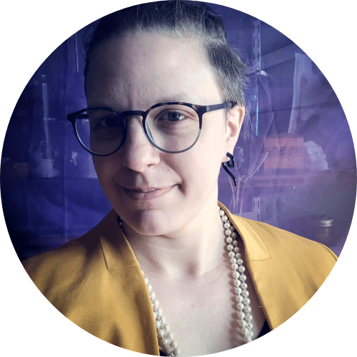 Photo of Juniper Caldera: wearing a canary yellow blazer and long, thick strand of pearls. They are wearing glasses with their hair tied on the top of their head with a close lipped smile.