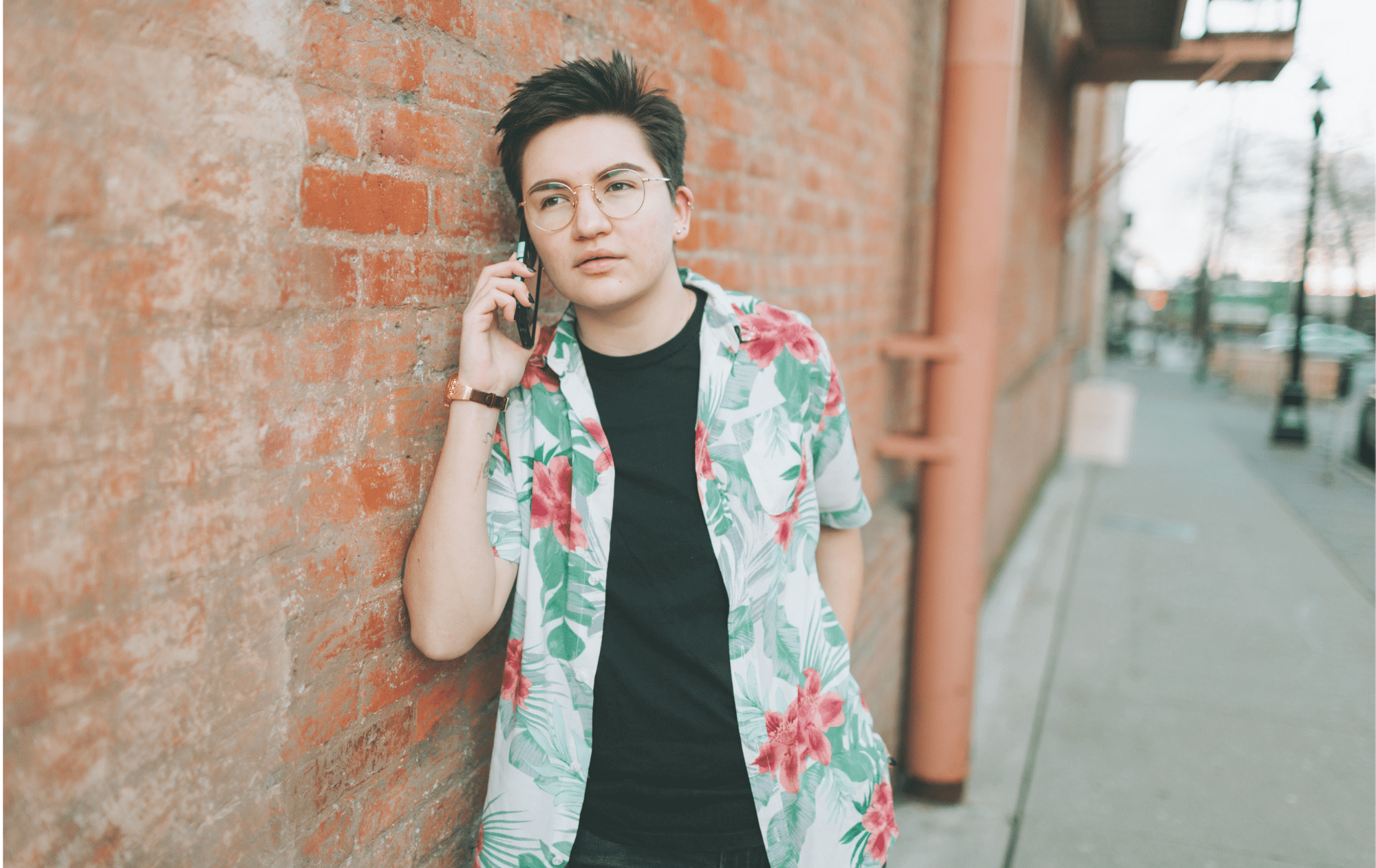 A non binary person leaning up against a brick building outside with a phone pressed up against their right ear. They are wearing gold, thin, wire classes with a floral button up shirt with a black shirt underneath. Their face indicates that they are intently listening while their gaze is softly focused on something in the distance off camera.